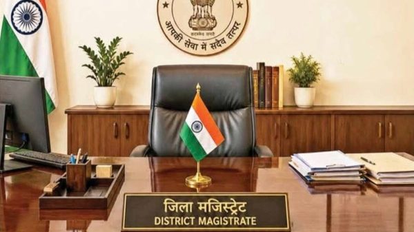 Front cover of a magazine showing a government office desk with a small Indian flag, a black leather chair, a wall emblem, and 'District Magistrate' nameplate; Hindi masthead at top.