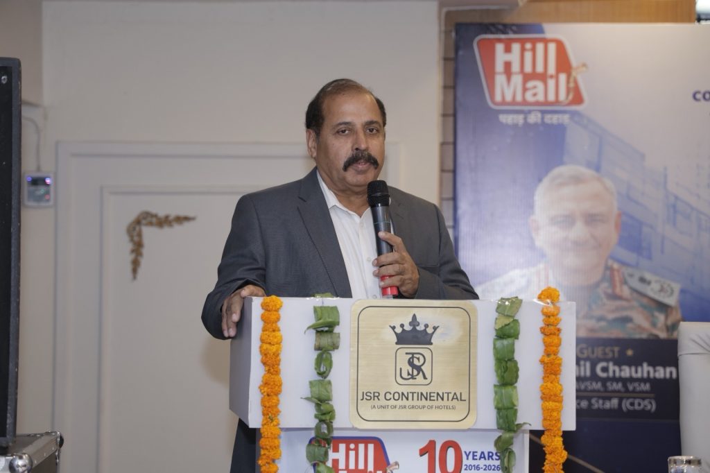 fbdc5ae9-2116-4c50-958e-1c58e25f5aea | No. 1 Indian Media News Portal Man in a gray suit speaks into a handheld microphone at a podium adorned with orange marigold garlands and green leaves.