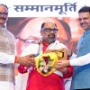 Three men on stage: central recipient in red with scarf receives a bouquet and plaque from two men in traditional Indian attire.