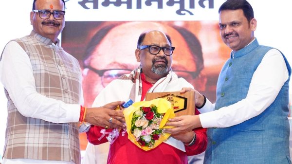 Three men on stage: central recipient in red with scarf receives a bouquet and plaque from two men in traditional Indian attire.