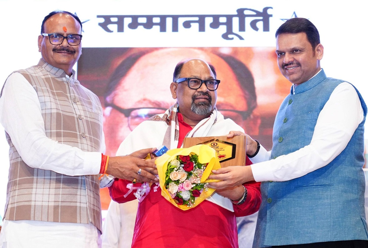 Three men on stage: central recipient in red with scarf receives a bouquet and plaque from two men in traditional Indian attire.