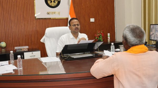A man in a white shirt sits at a large wooden desk in an office, speaking while handling documents as another person in an orange outfit sits across from him.