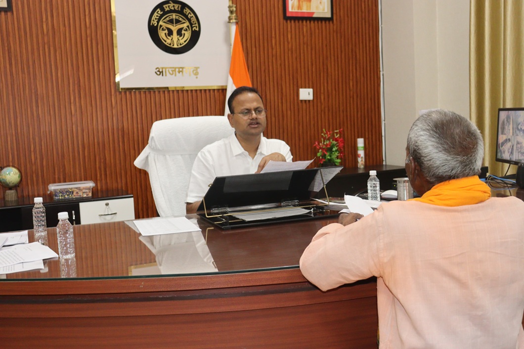A man in a white shirt sits at a large wooden desk in an office, speaking while handling documents as another person in an orange outfit sits across from him.