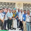 Group of men posing for a photo at an event; central man wears a garland and turban while others stand with badges in a room with patterned blinds and a clock behind them.