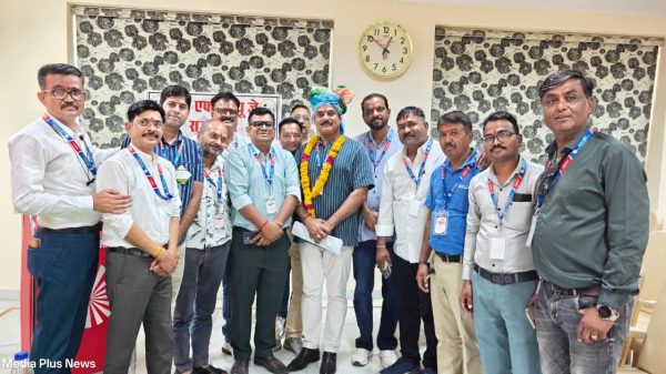 Group of men posing for a photo at an event; central man wears a garland and turban while others stand with badges in a room with patterned blinds and a clock behind them.