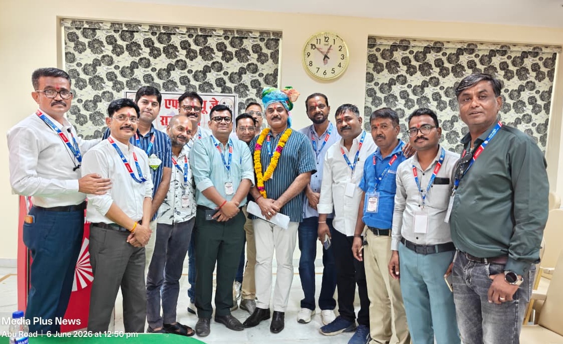 Group of men posing for a photo at an event; central man wears a garland and turban while others stand with badges in a room with patterned blinds and a clock behind them.