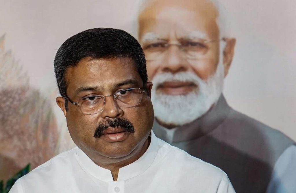 img_8792 | No. 1 Indian Media News Portal Man with mustache and glasses in a white shirt, speaking, with a large poster of an elderly man with a white beard in the background.