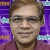 Portrait of a middle-aged man wearing glasses and a maroon shirt, standing in front of a purple backdrop with Hindi logos.
