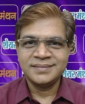 Portrait of a middle-aged man wearing glasses and a maroon shirt, standing in front of a purple backdrop with Hindi logos.