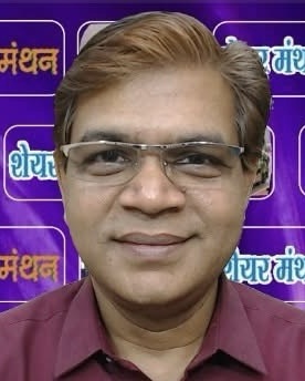 Portrait of a middle-aged man wearing glasses and a maroon shirt, standing in front of a purple backdrop with Hindi logos.