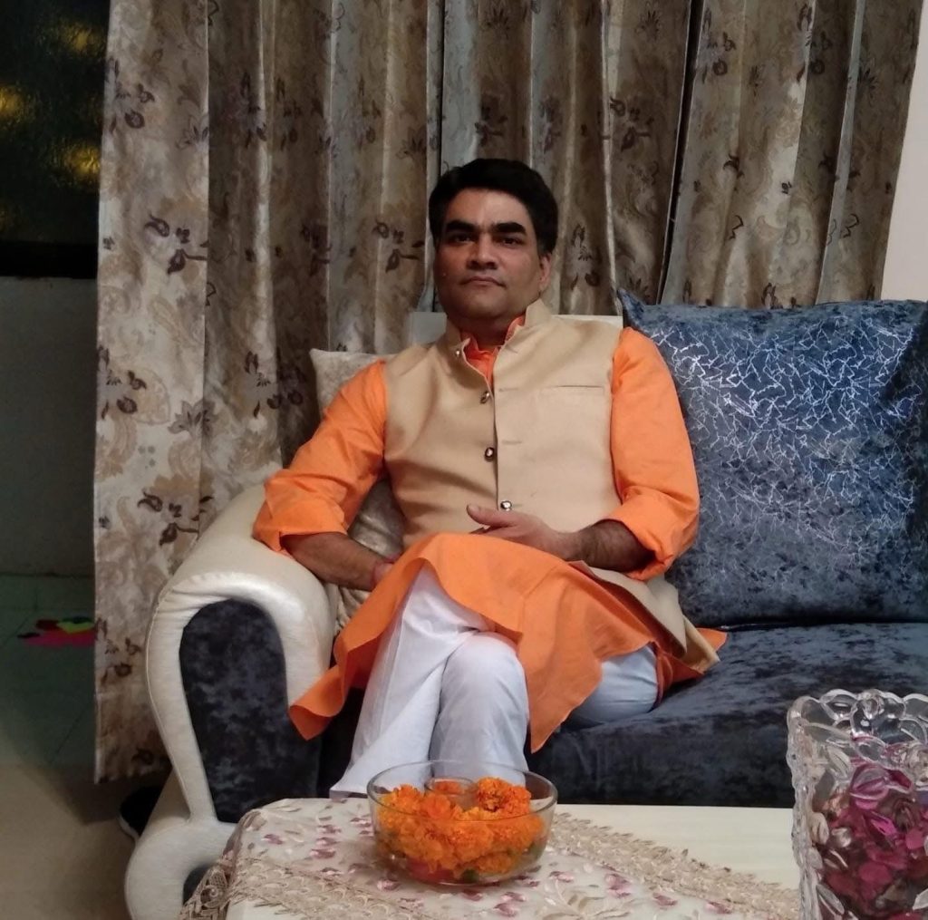 img_8907 | No. 1 Indian Media News Portal Man in an orange kurta and beige vest sitting on a sofa, looking at the camera in a living room with floral curtains behind him.