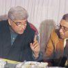 Two men sit at a dining table eating and talking, one gesturing with his hand while the other listens.