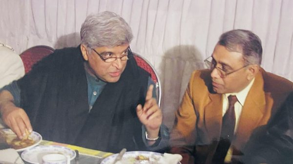Two men sit at a dining table eating and talking, one gesturing with his hand while the other listens.