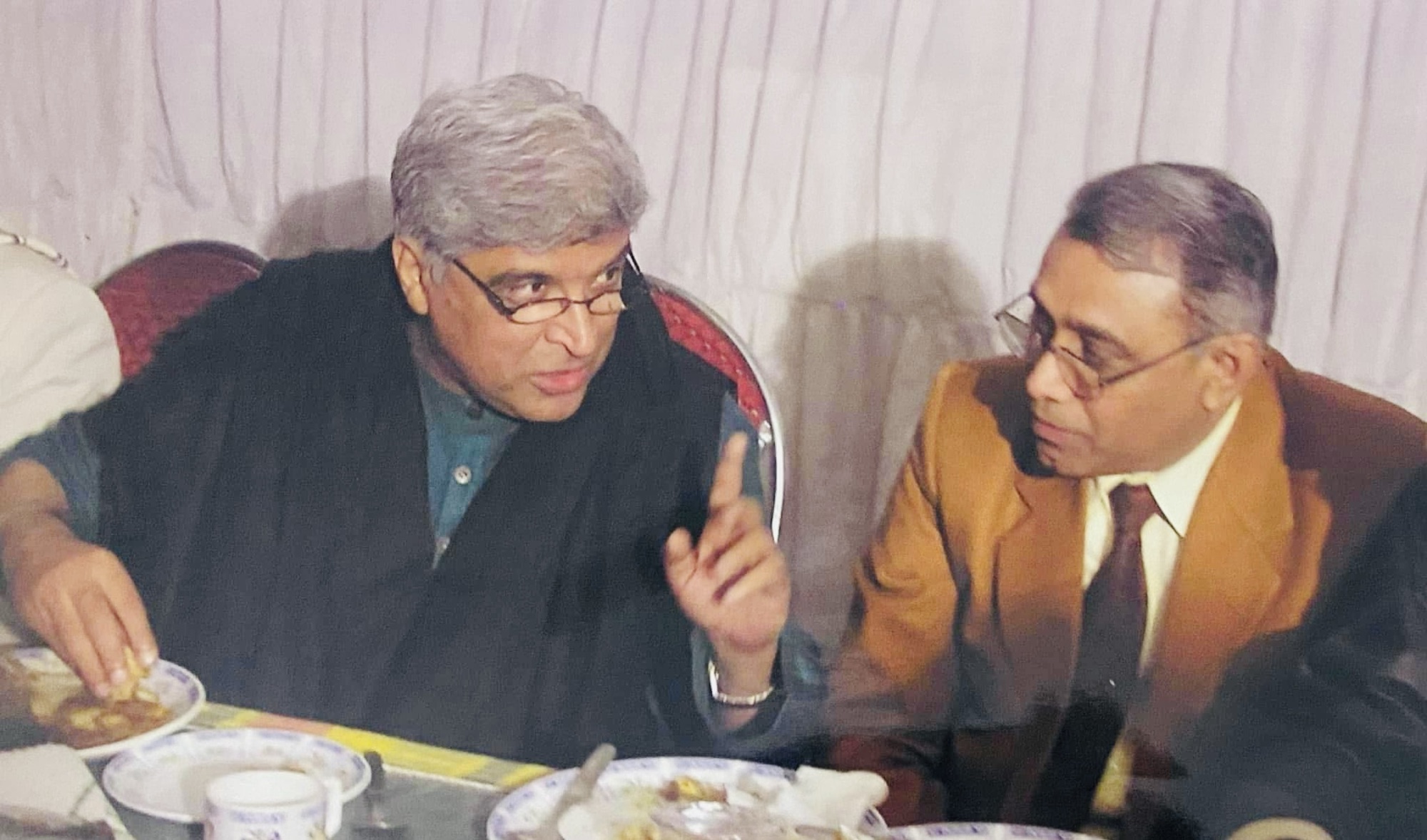 Two men sit at a dining table eating and talking, one gesturing with his hand while the other listens.