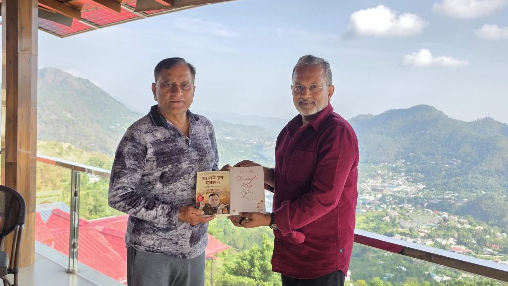 Two men stand on a balcony with a mountain view, exchanging two books between them (one Hindi title, one English).