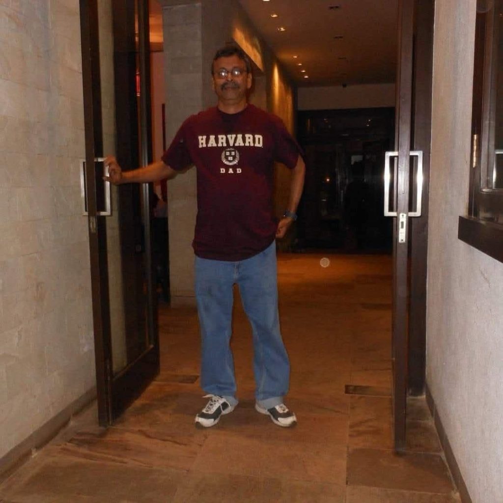 img_9126 | No. 1 Indian Media News Portal Man wearing a maroon Harvard Dad t-shirt, blue jeans, and sneakers stands in a doorway, one hand on the door frame in a dim hallway.