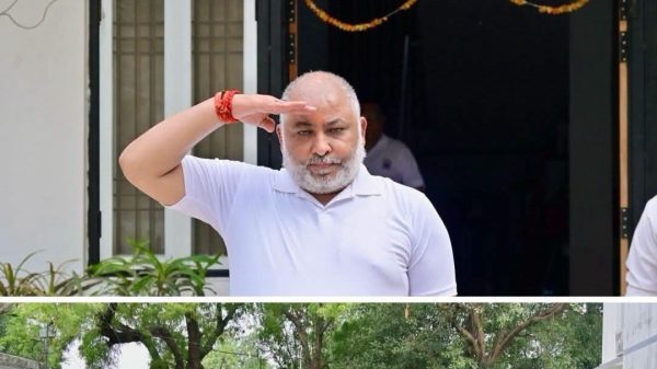 Bearded man in a white polo shirt saluting with his right hand raised, standing on a red platform; a line of uniformed guards with rifles stands at attention nearby, with a white SUV parked in the background.