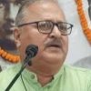 Man with glasses speaking at a microphone at a formal event, wearing a light green kurta.