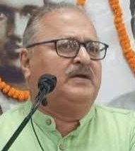 Man with glasses speaking at a microphone at a formal event, wearing a light green kurta.