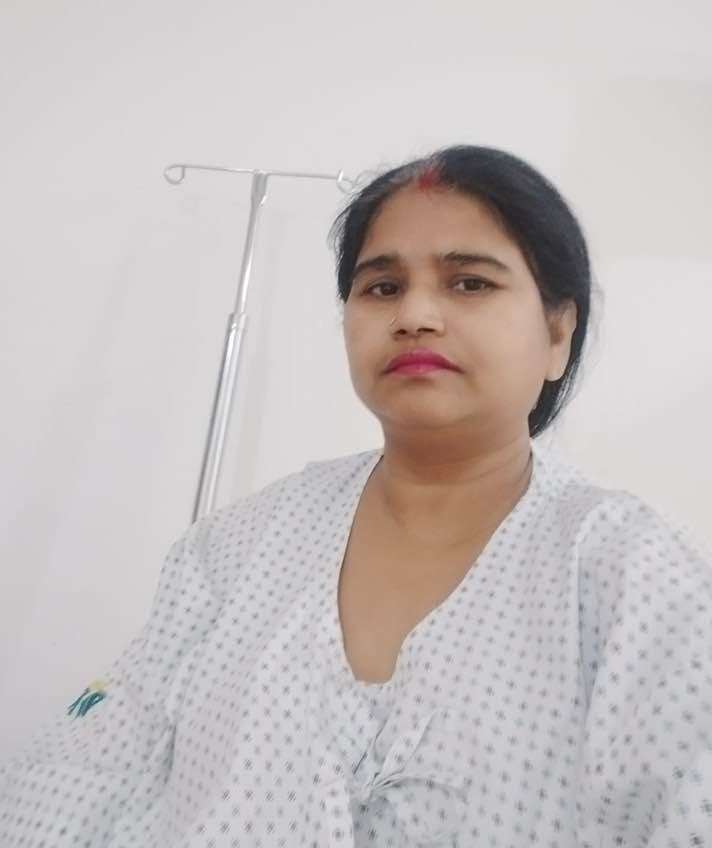 Woman in a patterned hospital gown seated in a clinical room with an IV stand behind her.