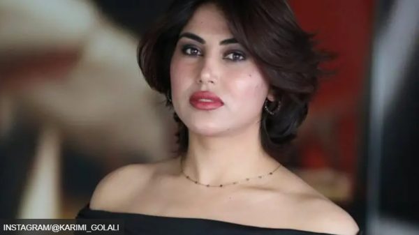 Woman with short dark hair, red lipstick, off‑the‑shoulder black top, delicate necklace, looking at the camera.