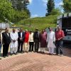Group of men in traditional and formal clothing pose outdoors beside a sign reading 'Doodh Ganga Resort and Restaurant' with a grassy hill and trees in the background.