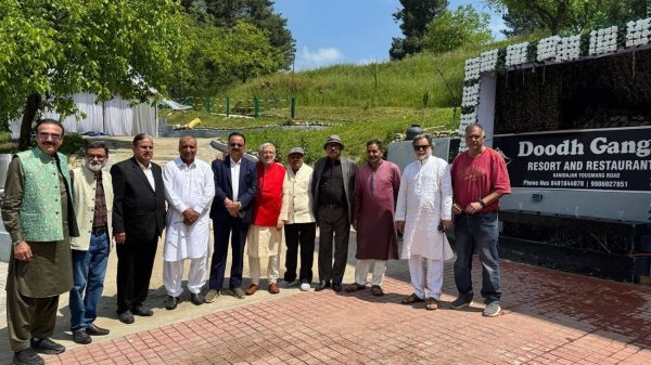 Group of men in traditional and formal clothing pose outdoors beside a sign reading 'Doodh Ganga Resort and Restaurant' with a grassy hill and trees in the background.