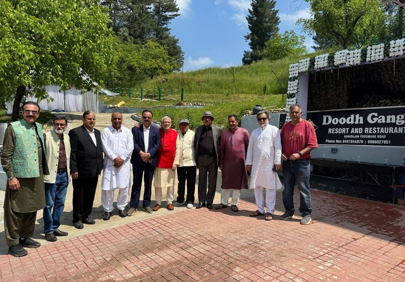 Group of men in traditional and formal clothing pose outdoors beside a sign reading 'Doodh Ganga Resort and Restaurant' with a grassy hill and trees in the background.