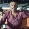 A man in a maroon shirt sits on a wooden bench outdoors at night, facing the camera.