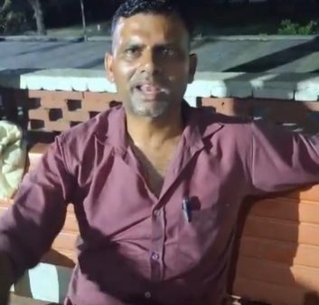 A man in a maroon shirt sits on a wooden bench outdoors at night, facing the camera.