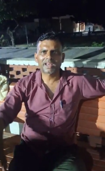 A man in a maroon shirt sits on a wooden bench outdoors at night, facing the camera.