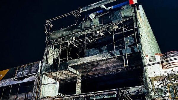 Nighttime view of a fire-damaged, scaffolding-covered building with a charred facade and yellow caution tape surrounding the site.