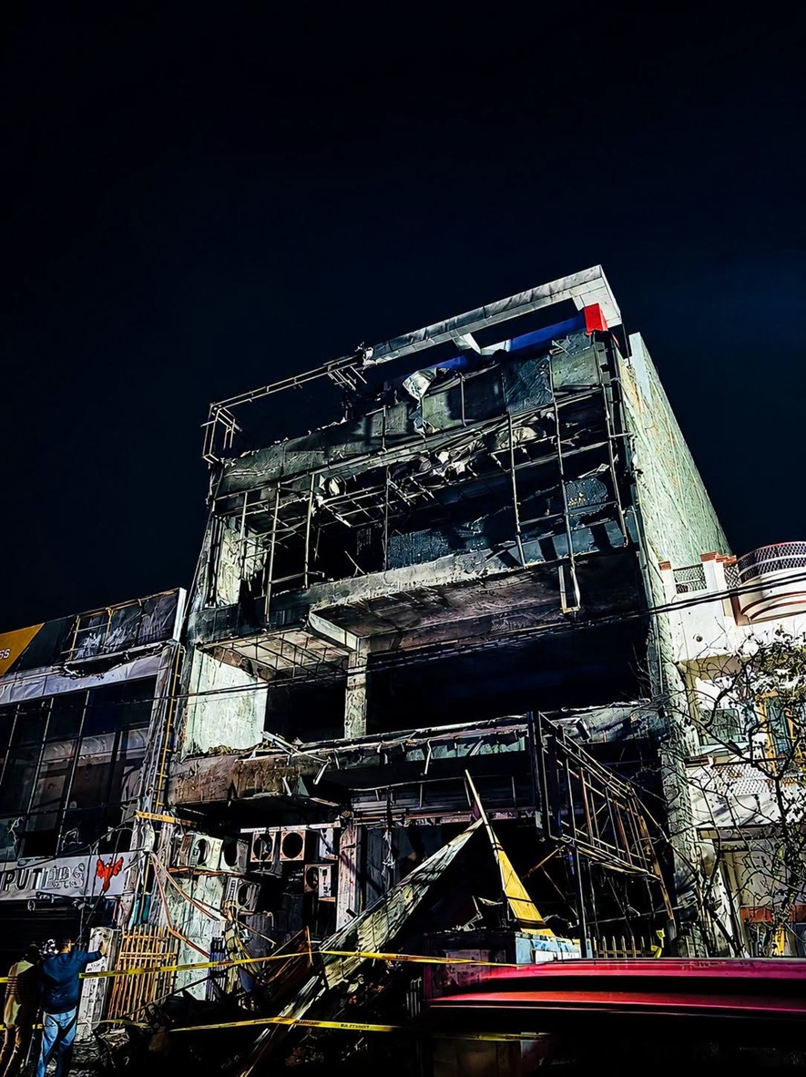 Nighttime view of a fire-damaged, scaffolding-covered building with a charred facade and yellow caution tape surrounding the site.