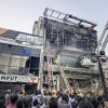 Crowd gathers as firefighters work at a partially collapsed multi-story building, with a crane and rescue ladders in use.