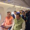 Two men sit in the front row of an airplane cabin: one wearing an orange vest, the other a pale green vest, with passengers behind them.