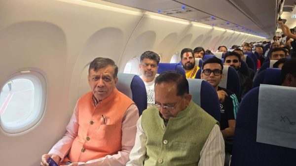 Two men sit in the front row of an airplane cabin: one wearing an orange vest, the other a pale green vest, with passengers behind them.