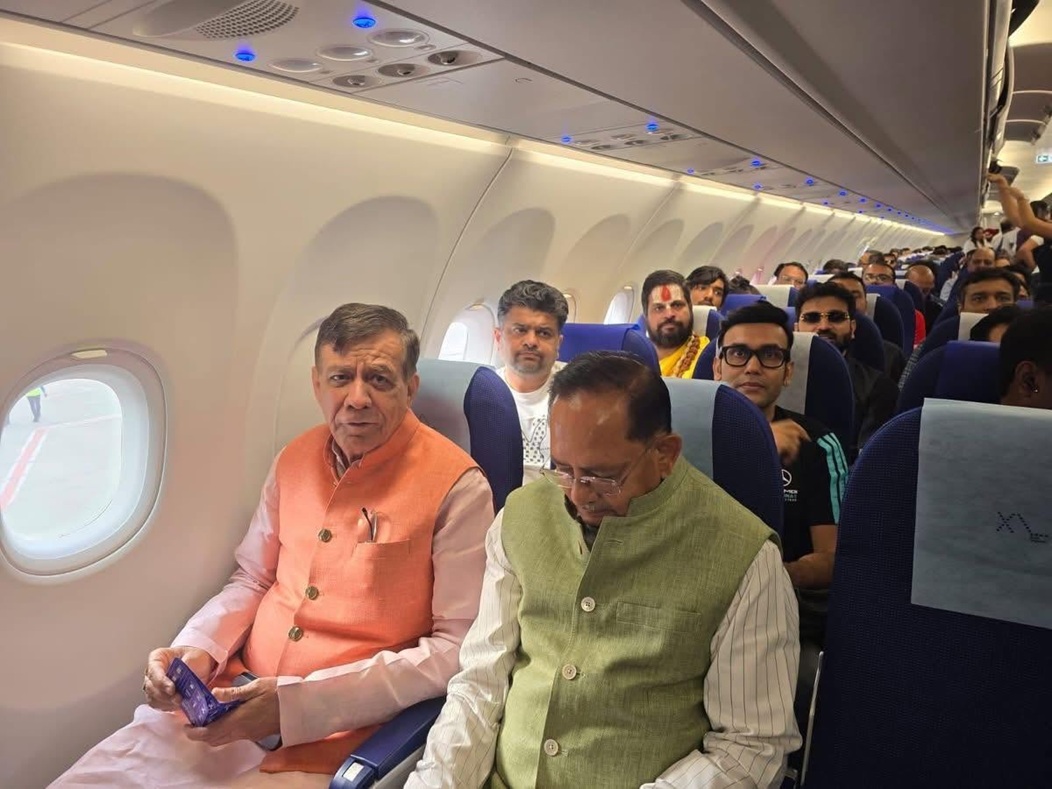 Two men sit in the front row of an airplane cabin: one wearing an orange vest, the other a pale green vest, with passengers behind them.