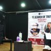 An elderly man sits on a bench on a dim stage, speaking into a microphone, with a large banner reading 'In Search of Truth' beside him.