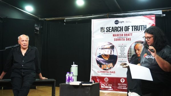 An elderly man sits on a bench on a dim stage, speaking into a microphone, with a large banner reading 'In Search of Truth' beside him.