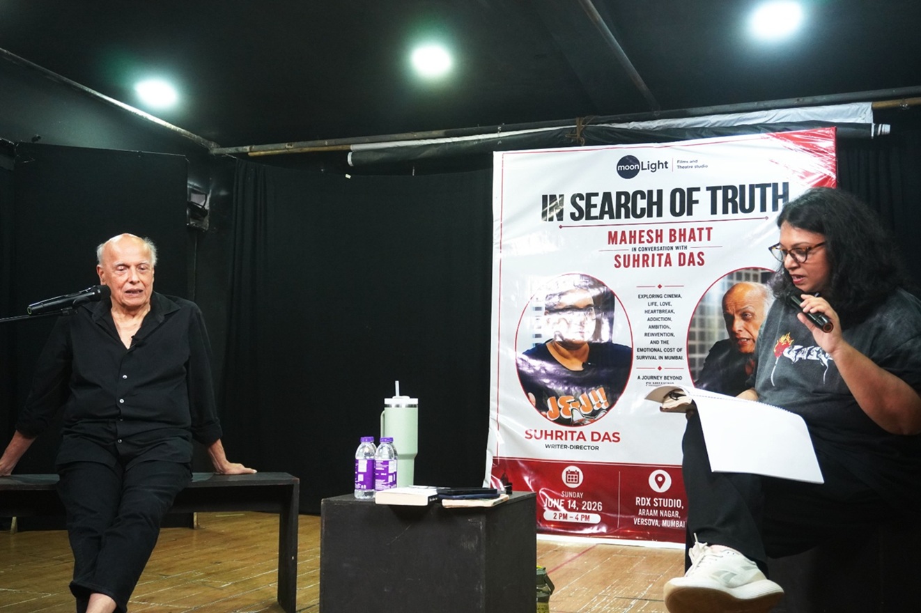 An elderly man sits on a bench on a dim stage, speaking into a microphone, with a large banner reading 'In Search of Truth' beside him.
