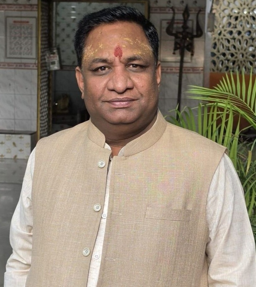 Portrait of a man wearing a beige Nehru jacket over a cream kurta, with a red tilak on his forehead, standing indoors with plants in the background.