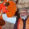Indian leader in white kurta and orange scarf raises a gilded ceremonial staff during a celebration with orange flowers in the background.