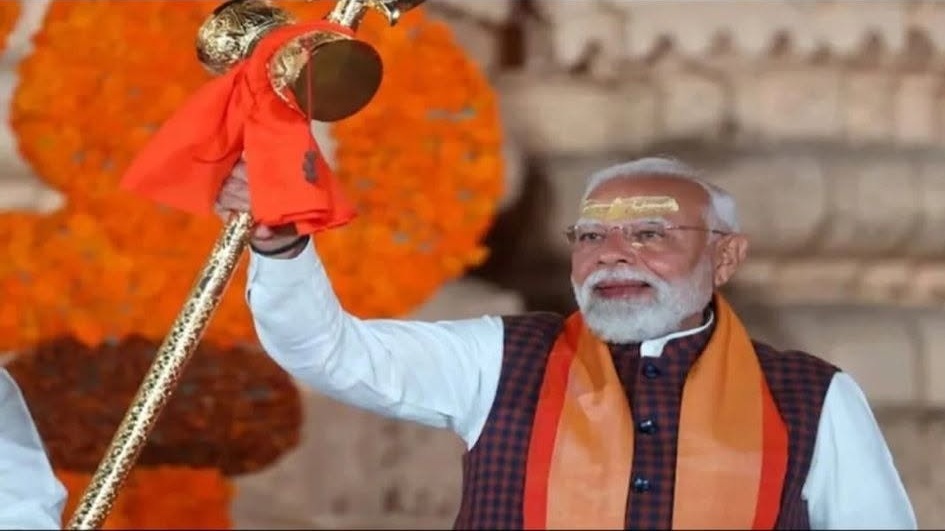 Indian leader in white kurta and orange scarf raises a gilded ceremonial staff during a celebration with orange flowers in the background.
