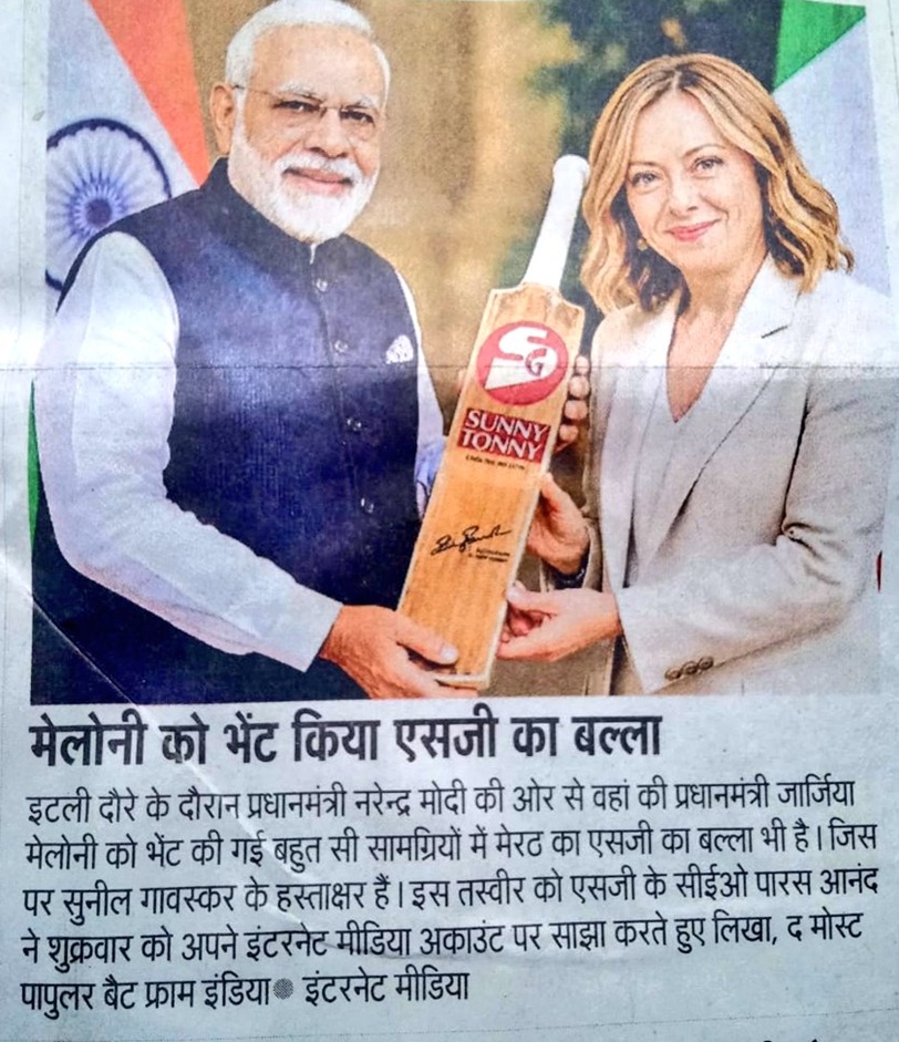 Prime Minister Narendra Modi and a smiling woman present a wooden cricket bat labeled 'Sunny Tonny' at a formal event, with a flag backdrop.