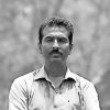 Man with a mustache wearing a light button-down shirt standing outdoors with a neutral expression, in a grayscale photo.