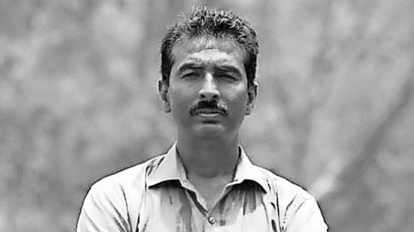 Man with a mustache wearing a light button-down shirt standing outdoors with a neutral expression, in a grayscale photo.