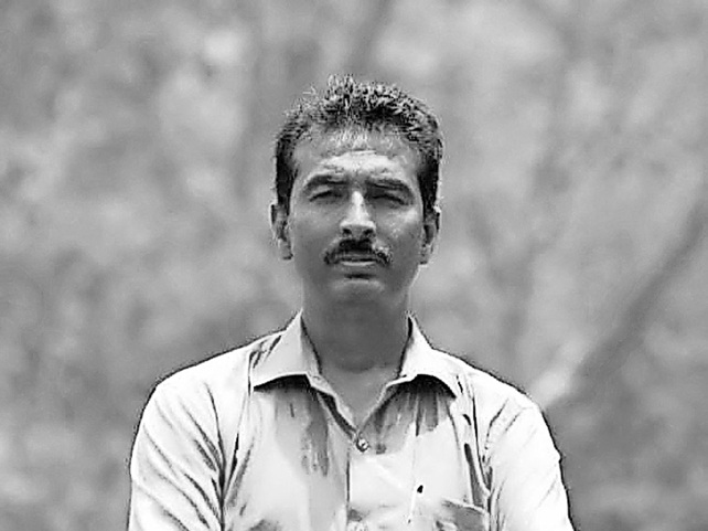 Man with a mustache wearing a light button-down shirt standing outdoors with a neutral expression, in a grayscale photo.