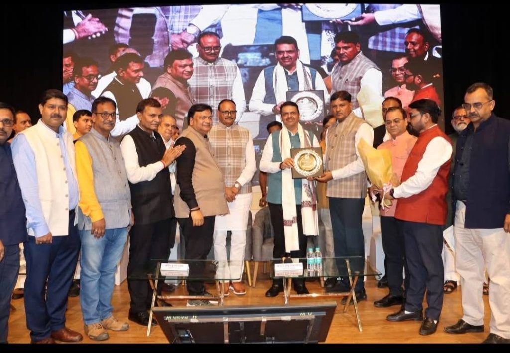mumbai ayojan | No. 1 Indian Media News Portal Group of men on a stage posing with plaques during an award ceremony; two center figures exchange a circular plaque while others applaud, a large screen backdrop behind.