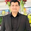 Portrait of a man in a black suit posing for a photo indoors, with red and yellow flowers in the background.