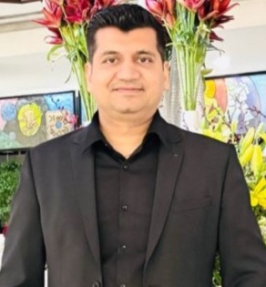 Portrait of a man in a black suit posing for a photo indoors, with red and yellow flowers in the background.